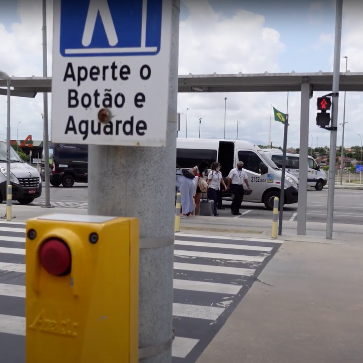 Poste de semáforo com botoeira amarela para pedestres em uma faixa de travessia. Uma placa acima da botoeira instrui "Aperte o Botão e Aguarde". Ao fundo, uma faixa de pedestres, vans estacionadas e um semáforo veicular com luz vermelha acesa.