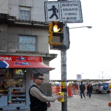 Homem, possivelmente com deficiência visual, usando óculos escuros e bengala longa, aciona a botoeira amarela de um semáforo para pedestres. O semáforo de pedestres acima exibe o sinal de "não atravesse" (mão espalmada na cor âmbar). Uma placa acima do semáforo instrui "PEDESTRE AGUARDE O SINAL VERDE".