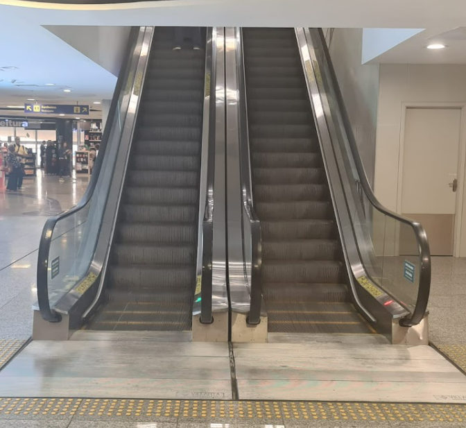 Escadas rolantes lado a lado em um ambiente de aeroporto, com sinalização tátil amarela no piso e corrimãos pretos de ambos os lados.
