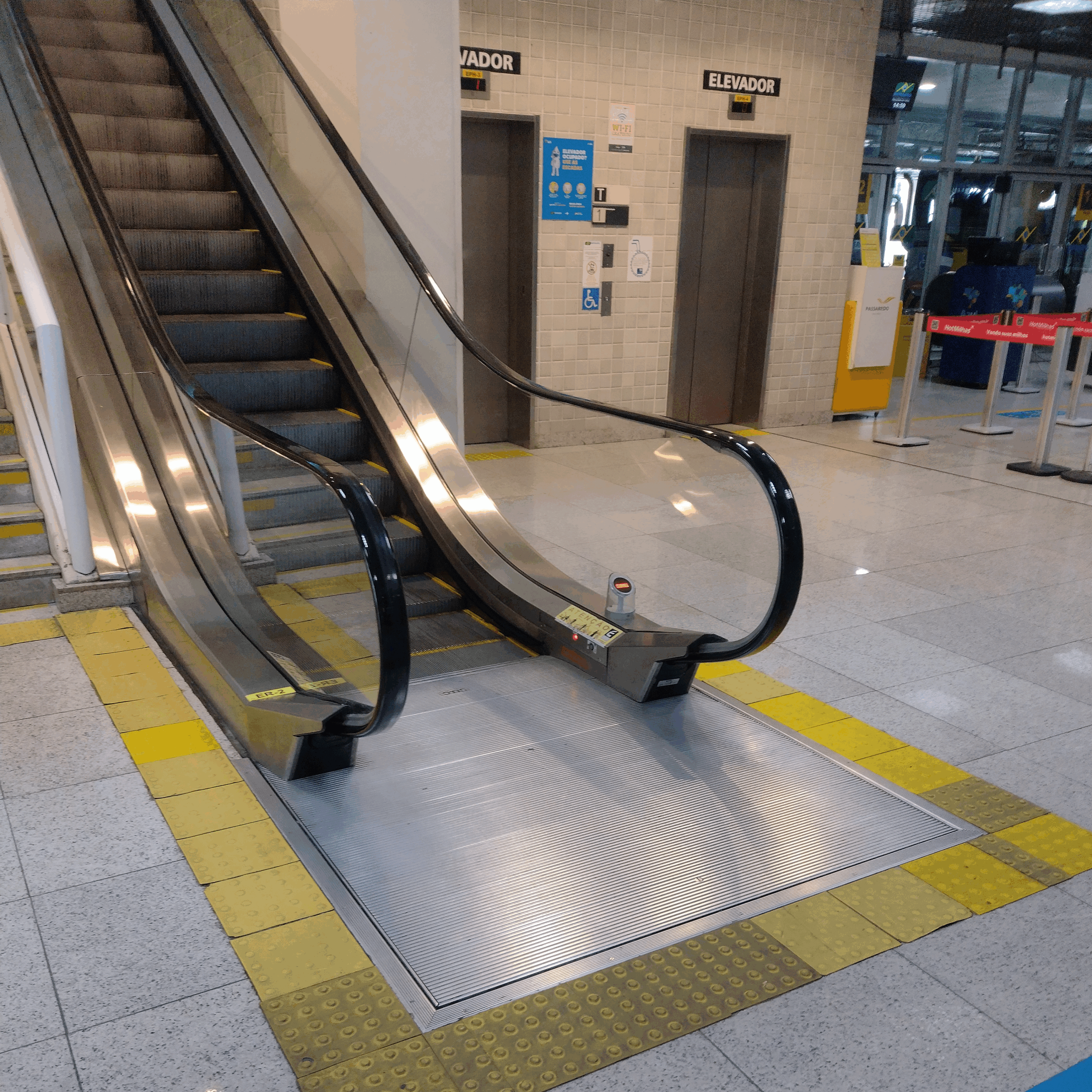 Escada rolante com piso tátil de alerta na área de entrada, localizada ao lado de elevadores e com demarcação em piso podotátil de cor amarela, evidenciando acessibilidade e segurança para pessoas com deficiência visual.