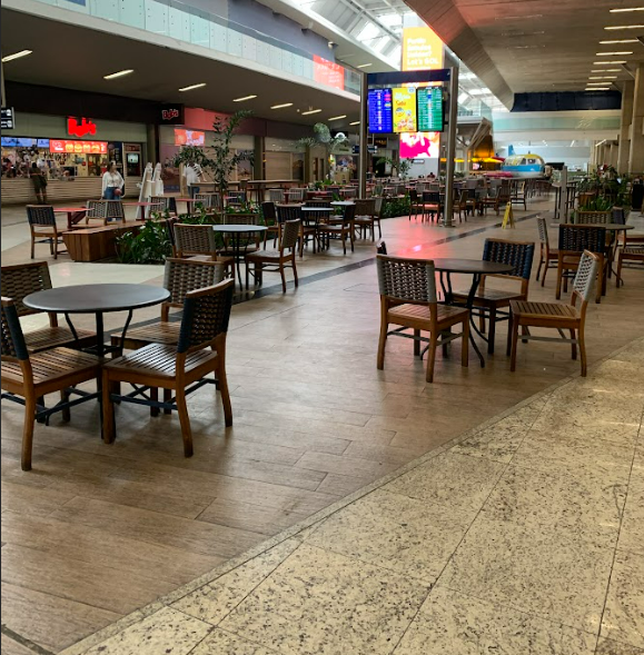 Vista ampla de uma praça de alimentação em um aeroporto, com mesas e cadeiras distribuídas de forma espaçada, permitindo a circulação de pessoas com deficiência ou mobilidade reduzida. Ao fundo, há estabelecimentos comerciais e um painel eletrônico com informações de voos. A circulação é facilitada por corredores largos e livres de obstáculos.